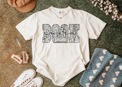 White t-shirt with 'Book Worm' design on a textured surface with accessories.