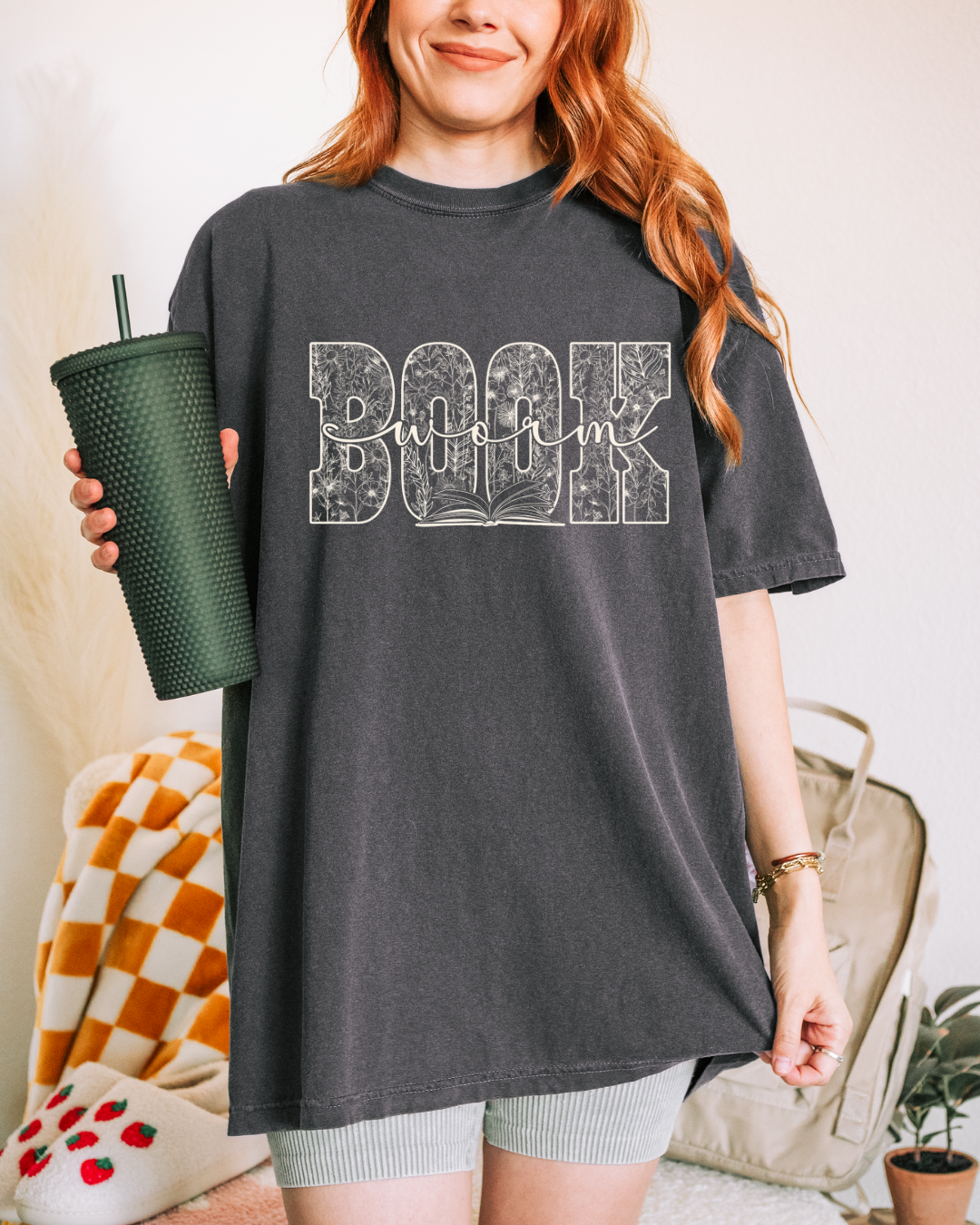 Person wearing a dark gray t-shirt with 'Book Worm' design, holding a green tumbler.