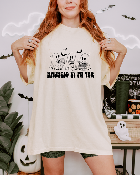 A woman wearing a cream-colored oversized t-shirt with a black graphic of three ghosts and the text 'HAUNTED BY MY TBR'. The background is decorated for Halloween with a pumpkin, plants, and a shelf.