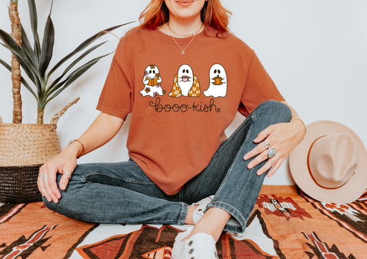 A woman sitting cross-legged on a patterned rug wearing an oversized rust-orange graphic T-shirt with three cartoon ghosts and the text 'boo-kish', paired with blue jeans and white sneakers. A straw hat and potted plant are beside her against a light wall.