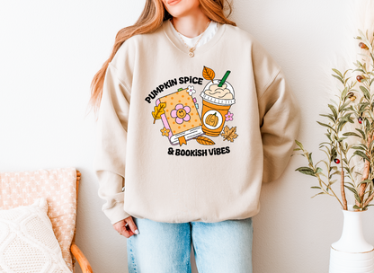 A woman wearing a beige crewneck sweatshirt with a colorful graphic that reads 'PUMPKIN SPICE & BOOKISH VIBES' alongside a pumpkin, iced coffee, and autumn leaves. She is wearing light blue jeans and standing in a brightly lit room with a chair and potted plant.
