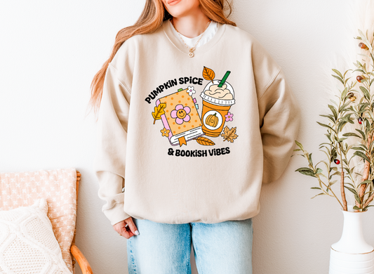 A woman wearing a beige crewneck sweatshirt with a colorful graphic that reads 'PUMPKIN SPICE & BOOKISH VIBES' alongside a pumpkin, iced coffee, and autumn leaves. She is wearing light blue jeans and standing in a brightly lit room with a chair and potted plant.