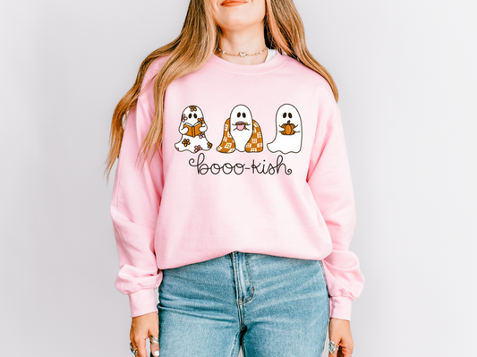 A woman wearing a light pink crewneck sweatshirt with a graphic of three cute cartoon ghosts and the text 'booo-kish'. She is also wearing blue jeans, set against a plain light gray background.