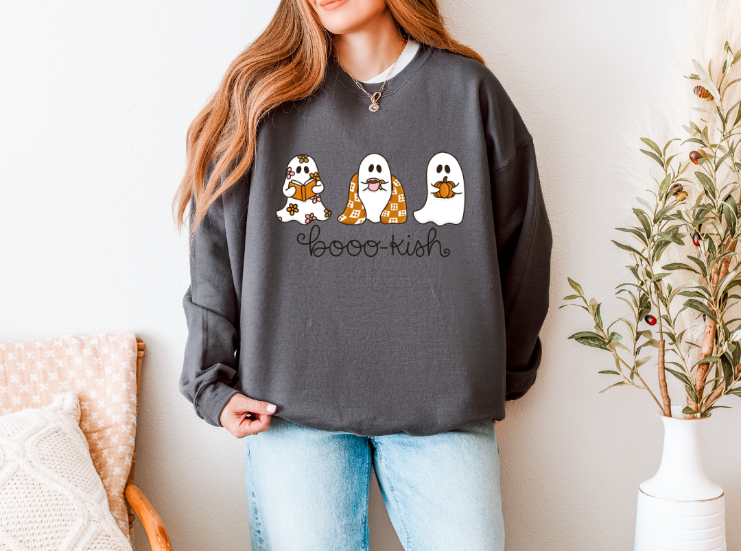A woman wearing a dark gray crewneck sweatshirt with a 'Boo-Ku-sh' graphic featuring three cute cartoon ghosts, paired with light blue jeans, in a brightly lit indoor setting.