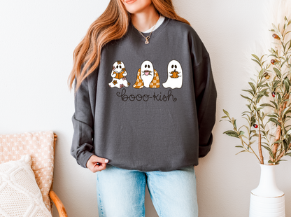 A woman wearing a dark gray crewneck sweatshirt with a 'Boo-Ku-sh' graphic featuring three cute cartoon ghosts, paired with light blue jeans, in a brightly lit indoor setting.