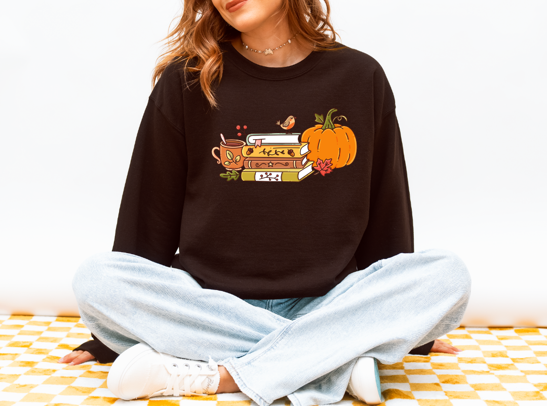 A woman sitting cross-legged wearing a dark brown crewneck sweatshirt with a colorful autumn graphic of a book, pumpkin, and bird, paired with light blue jeans and white sneakers.