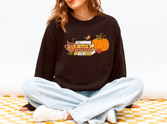 A woman sitting cross-legged wearing a dark brown crewneck sweatshirt with a colorful autumn graphic of a book, pumpkin, and bird, paired with light blue jeans and white sneakers.