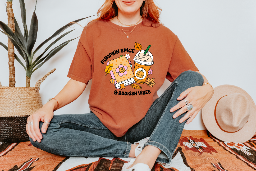 A woman sitting cross-legged on a patterned rug wearing an oversized rust-orange graphic T-shirt with a pumpkin spice latte illustration and the text 'Pumpkin Spice' and 'In Boobooth Vibes.' Jeans, white sneakers, and a straw hat are also visible in a styled indoor setting with a plant and wall.