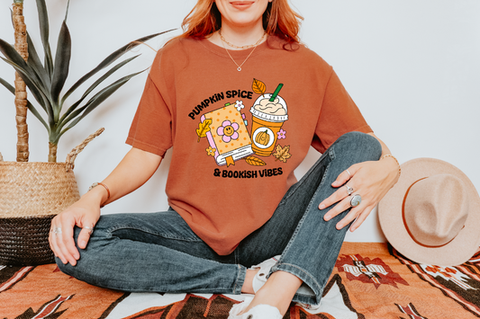 A woman sitting cross-legged on a patterned rug wearing an oversized rust-orange graphic T-shirt with a pumpkin spice latte illustration and the text 'Pumpkin Spice' and 'In Boobooth Vibes.' Jeans, white sneakers, and a straw hat are also visible in a styled indoor setting with a plant and wall.