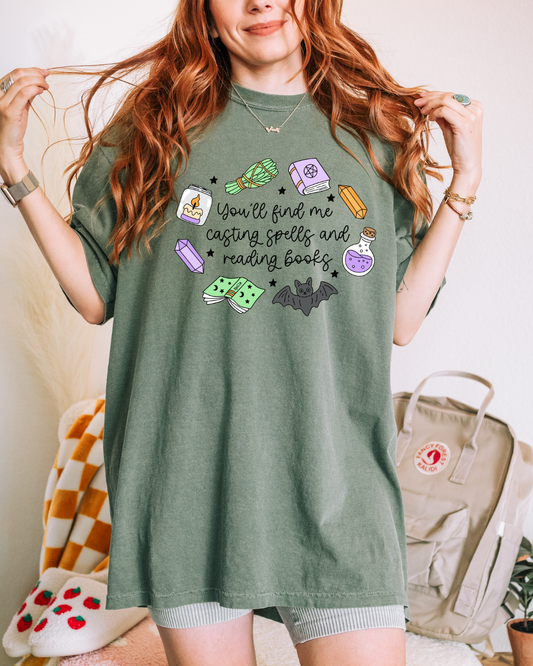 A woman wearing an oversized sage green graphic t-shirt with a witchy reading theme, featuring illustrations of a purple potion bottle, green potion jar, book, bat, and spells, with the text 'You'll find me casting spells and reading books'.